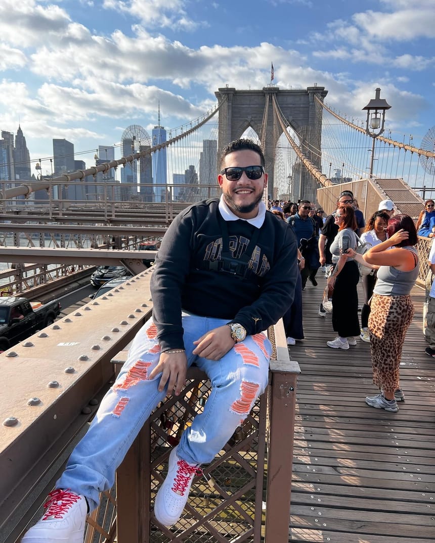 Cristian Rodríguez sentado en la barandilla del Puente de Brooklyn, sonriendo con gafas de sol